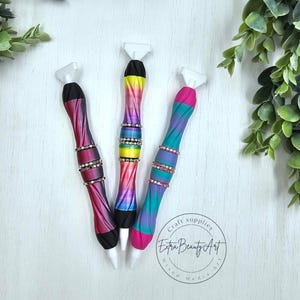 May include: Three colorful craft tools with white tips and jewel accents. The handles feature vibrant gradients of pink, purple, blue, and yellow. The tools are labeled "Extra Beauty Art" and are surrounded by greenery.