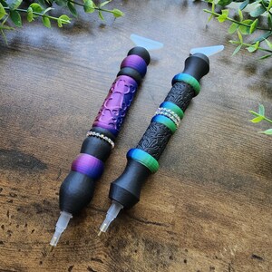 May include: Two diamond painting pens with black bodies and colorful accents. One pen has a purple, textured grip with a silver band, while the other has a black, patterned grip with a silver band. Both pens have clear tips and white silicone attachments.