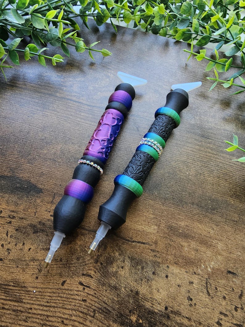 May include: Two diamond painting pens with black bodies and colorful accents. One pen has a purple, textured grip with a silver band, while the other has a black, patterned grip with a silver band. Both pens have clear tips and white silicone attachments.
