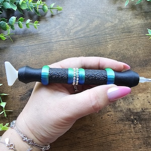 May include: Black diamond painting pen with a white tip and a patterned grip. The pen is decorated with green and blue rings, and a silver ring with clear stones. A crafting tool for diamond painting.