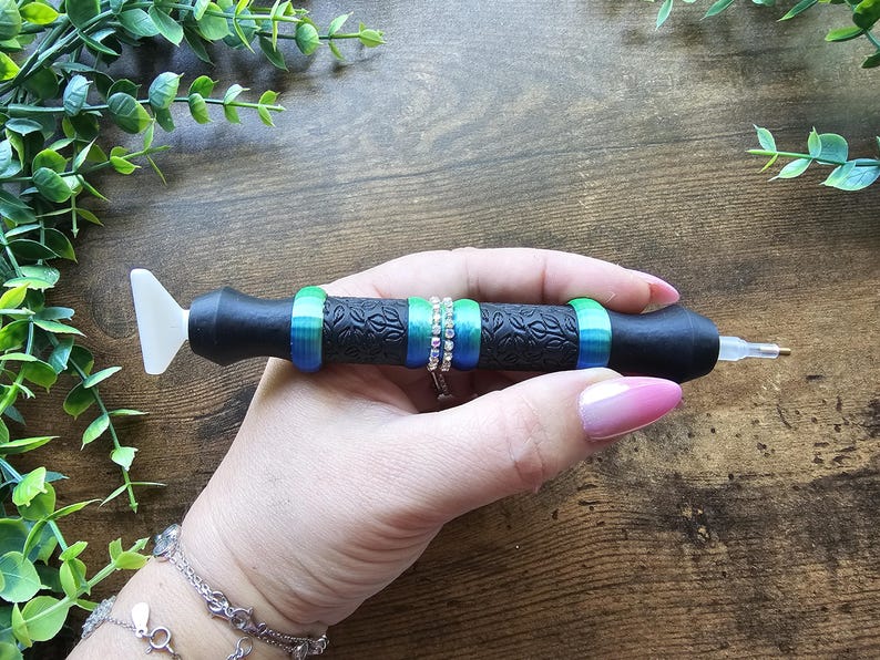 May include: Black diamond painting pen with a white tip and a patterned grip. The pen is decorated with green and blue rings, and a silver ring with clear stones. A crafting tool for diamond painting.