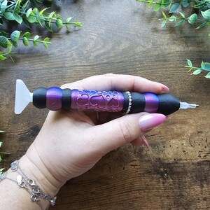 May include: A diamond painting pen with a black, purple, and silver design. The pen has a white tip and a decorative barrel with a heart pattern. The pen is held in a hand, with a wooden background and greenery.