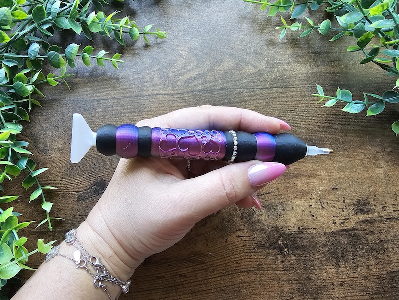 May include: A diamond painting pen with a black, purple, and silver design. The pen has a white tip and a decorative barrel with a heart pattern. The pen is held in a hand, with a wooden background and greenery.