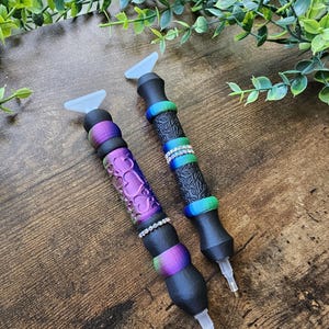 May include: Two diamond painting pens with black barrels and colorful accents. One pen has a purple heart design, the other has a black floral design. Both pens have clear tips and light blue paddle-shaped tops.