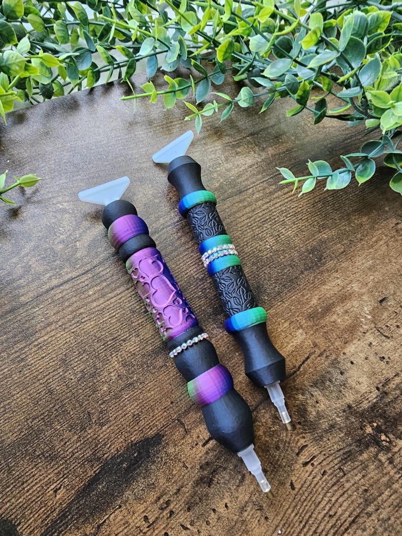May include: Two diamond painting pens with black barrels and colorful accents. One pen has a purple heart design, the other has a black floral design. Both pens have clear tips and light blue paddle-shaped tops.