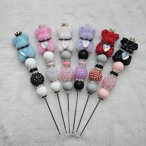 May include: A collection of six beaded skewers, each featuring a teddy bear charm with a crown and heart. The bears are in various colors: blue, pink, white, purple, red, and black. The skewers have decorative beads and are set against a white background.