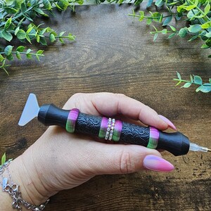 May include: Black diamond painting pen with a textured handle, accented with green and purple rings and a row of small, clear rhinestones. The pen has a clear, angled tip and a small metal tip. The pen is held in a hand with a pink and purple ombre manicure.