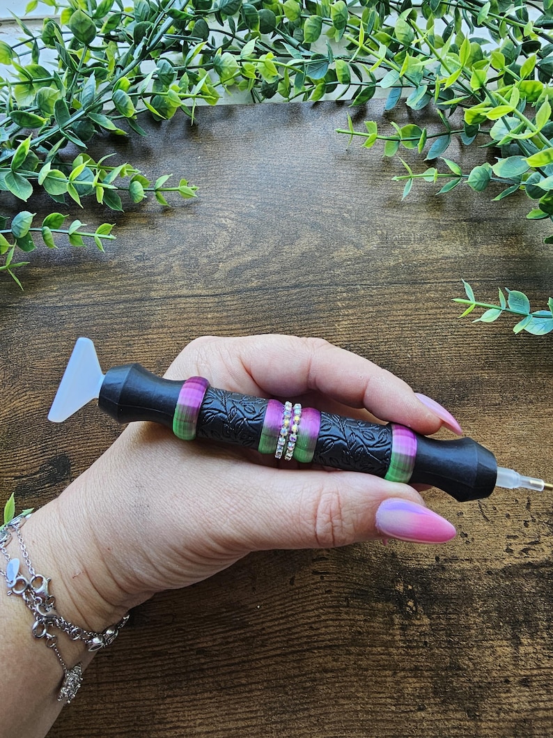 May include: Black diamond painting pen with a textured handle, accented with green and purple rings and a row of small, clear rhinestones. The pen has a clear, angled tip and a small metal tip. The pen is held in a hand with a pink and purple ombre manicure.