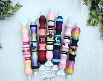 May include: A collection of colorful diamond painting pens with decorative handles. The pens feature various designs, including butterflies, bees, and rhinestones. The pens are arranged on a white surface, with green foliage in the background. The text "Extra Beauty Art" is visible.