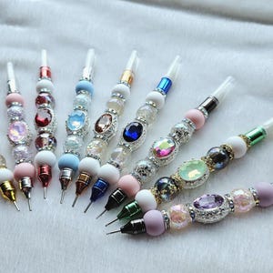 May include: A collection of beaded accessories with decorative tips. The beads are in various colors, including white, pink, blue, and green, and are accented with rhinestones and other embellishments. The tips are gold, silver, and black.