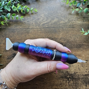 May include: A diamond painting pen with a black, purple, and blue ombre design. The pen features a heart pattern and a rhinestone band. The pen has a white tip and is held by a person.