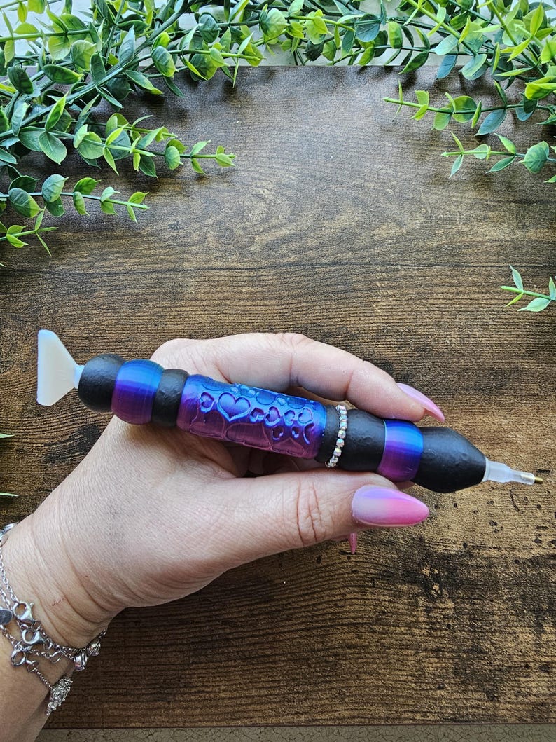 May include: A diamond painting pen with a black, purple, and blue ombre design. The pen features a heart pattern and a rhinestone band. The pen has a white tip and is held by a person.
