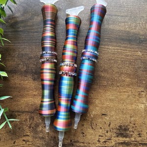 May include: Three colorful diamond painting pens with a rainbow striped design. Each pen has a clear, angled tip and a decorative band of rhinestones. The pens are displayed on a wooden surface.