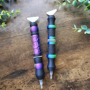 May include: Two diamond painting pens with black bodies and white tips. One pen has a purple heart design, the other has a textured pattern. Both pens have colorful rings and rhinestone accents.