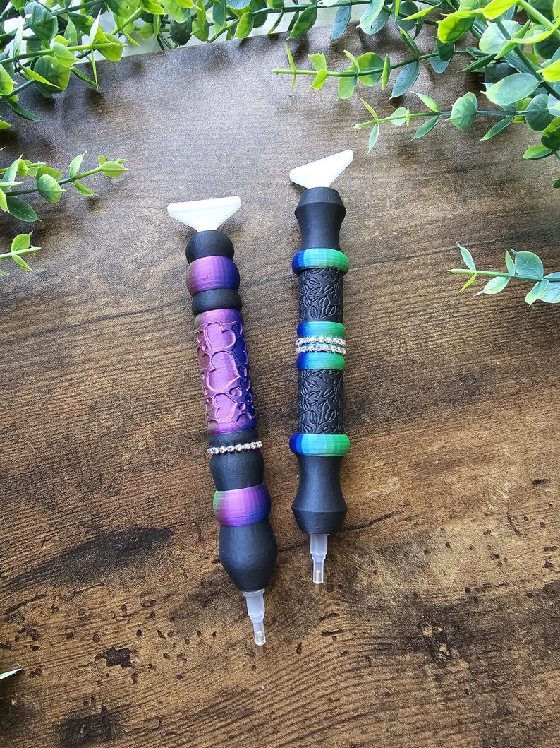 May include: Two diamond painting pens with black bodies and white tips. One pen has a purple heart design, the other has a textured pattern. Both pens have colorful rings and rhinestone accents.