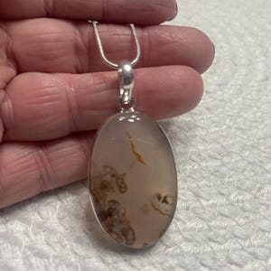 May include: Oval pendant necklace with a silver chain. The pendant features a translucent, light-coloured stone with brown and tan inclusions. The stone is set in a silver bezel.