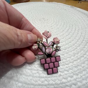 May include: A vintage floral brooch featuring a bouquet of pink roses and small white flowers in a pink pot. The brooch has a dark metal frame and is held by a hand. A charming accessory for a jacket or scarf.