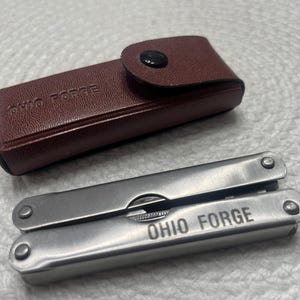 以下が含まれることがあります： 「OHIO FORGE」の文字が刻まれたシルバーのマルチツールと、同じ文字が刻まれた茶色の革製ケース。マルチツールには、折りたたまれた複数のツールがあります。ケースはスナップ留めです。