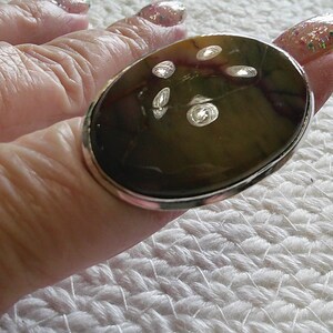 May include: An oval-shaped ring with a large, polished stone. The stone has a mix of brown and green hues with several clear, reflective inclusions. The ring has a silver-colored metal band.