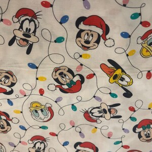 Könnte beinhalten: Weißer Stoff mit einem sich wiederholenden Muster von Disney-Figuren mit Weihnachtsmützen, darunter Micky Maus, Donald Duck und Goofy. Das Design umfasst auch bunte Weihnachtsbeleuchtung.