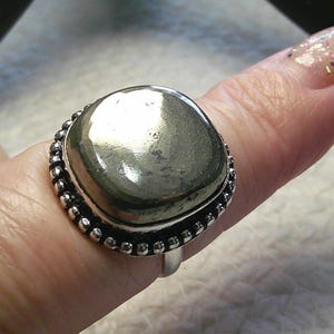 Puede incluir: Un anillo de plata con una gran piedra de pirita cuadrada. La pirita tiene una superficie metálica y reflectante. La banda del anillo es plateada y la piedra está rodeada por un borde de cuentas. El anillo se muestra en un dedo.