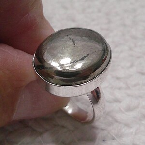 May include: A silver-toned ring featuring a large, oval-shaped, metallic pyrite gemstone set in a smooth bezel. The stone has a reflective, gunmetal-gray surface with natural inclusions and a polished finish, mounted on a simple, solid metal band.