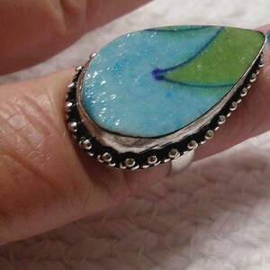May include: A teardrop-shaped ring featuring a textured, light blue and green surface with a dark blue line accent. The stone is set in a silver-toned metal bezel with a decorative beaded edge, mounted on a simple, smooth silver-toned metal band.