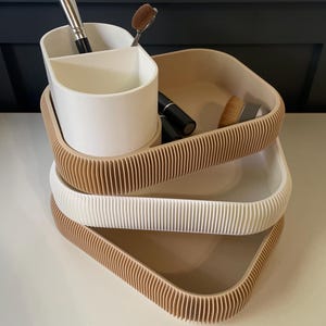May include: A three-tiered makeup organizer with a white divided cup and three trays in beige and white. The trays have a ribbed edge design. Makeup brushes and lipstick are visible.