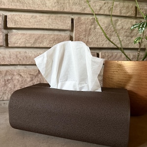 Puede incluir: Un dispensador de pañuelos marrón con una superficie texturizada, que contiene pañuelos blancos. El dispensador es rectangular con bordes redondeados. Una planta en una maceta de madera es visible en el fondo.