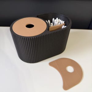Puede incluir: Un contenedor negro, acanalado, impreso en 3D con una tapa circular y una tapa curva separada. El contenedor contiene bastoncillos de algodón y una herramienta de madera. Las tapas son de color marrón claro.
