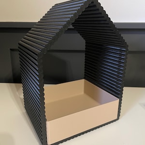 Puede incluir: Un contenedor de almacenamiento decorativo negro en forma de casa. El exterior está hecho de varillas negras redondeadas y paralelas. El interior presenta una bandeja de color beige claro. El contenedor está diseñado para colocarse sobre una superficie plana.
