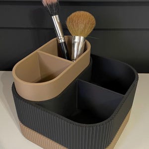 Pode incluir: Um organizador de maquilhagem de dois níveis, preto e bege. A camada superior bege contém dois pincéis de maquilhagem. A camada inferior é preta com detalhes canelados verticais e uma base bege. Projetado para guardar pincéis e outros produtos de beleza.