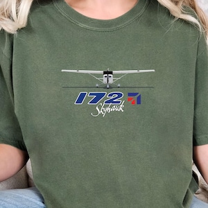 Puede incluir: Una camiseta de manga corta color verde oliva con un gráfico de un avión Cessna 172 Skyhawk blanco. Debajo del avión, el texto dice 172 en una fuente azul grande, seguido de la palabra Skyhawk en letra cursiva blanca y un pequeño logotipo rojo y azul.