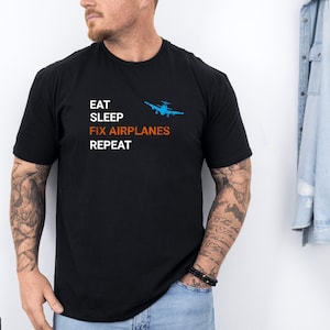 Puede incluir: Camiseta negra con el texto "EAT SLEEP FIX AIRPLANES REPEAT" en blanco y naranja. Un gráfico de avión azul está encima de las palabras "FIX AIRPLANES". La camiseta es de manga corta.