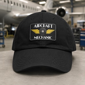 Puede incluir: Una gorra de béisbol negra con un parche rectangular en la parte delantera. El parche muestra el texto AIRCRAFT MECHANIC en letras blancas, centrado encima y debajo de un gráfico de una turbina de motor a reacción flanqueado por dos alas amarillas.