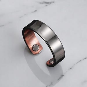 Op de afbeelding: Een zilverkleurige manchetarmband met een koperen binnenkant en een magnetische sluiting. De armband heeft een plat, rechthoekig ontwerp en wordt gepresenteerd op een wit marmeren oppervlak. De manchet is een modern sieraad.