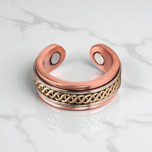May include: A copper-colored magnetic ring with a decorative gold and silver band. The ring has two magnets on the open ends. The ring is on a white marble surface.