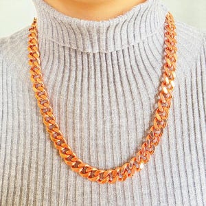 Puede incluir: Un collar de cadena de color cobre con eslabones grandes e entrelazados. El collar se lleva sobre un jersey de cuello alto gris. La cadena tiene un acabado metálico brillante y es una pieza llamativa.