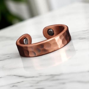 May include: Copper-colored cuff bracelet with a hammered texture. The open-ended design features two small, dark magnets. The bracelet is displayed on a white marble surface.