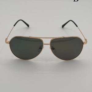 Puede incluir: Gafas de sol de aviador con montura dorada y lentes oscuras. Las gafas tienen un doble puente y patillas negras. La letra "B" es visible en la esquina superior derecha. Dise&ntilde;adas para protecci&oacute;n ocular y moda.