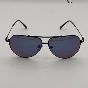 Puede incluir: Gafas de sol de aviador negras con lentes tintadas de p&uacute;rpura. Las gafas de sol tienen una forma cl&aacute;sica de l&aacute;grima y una montura de metal negro. La letra "C" es visible en la esquina superior derecha.
