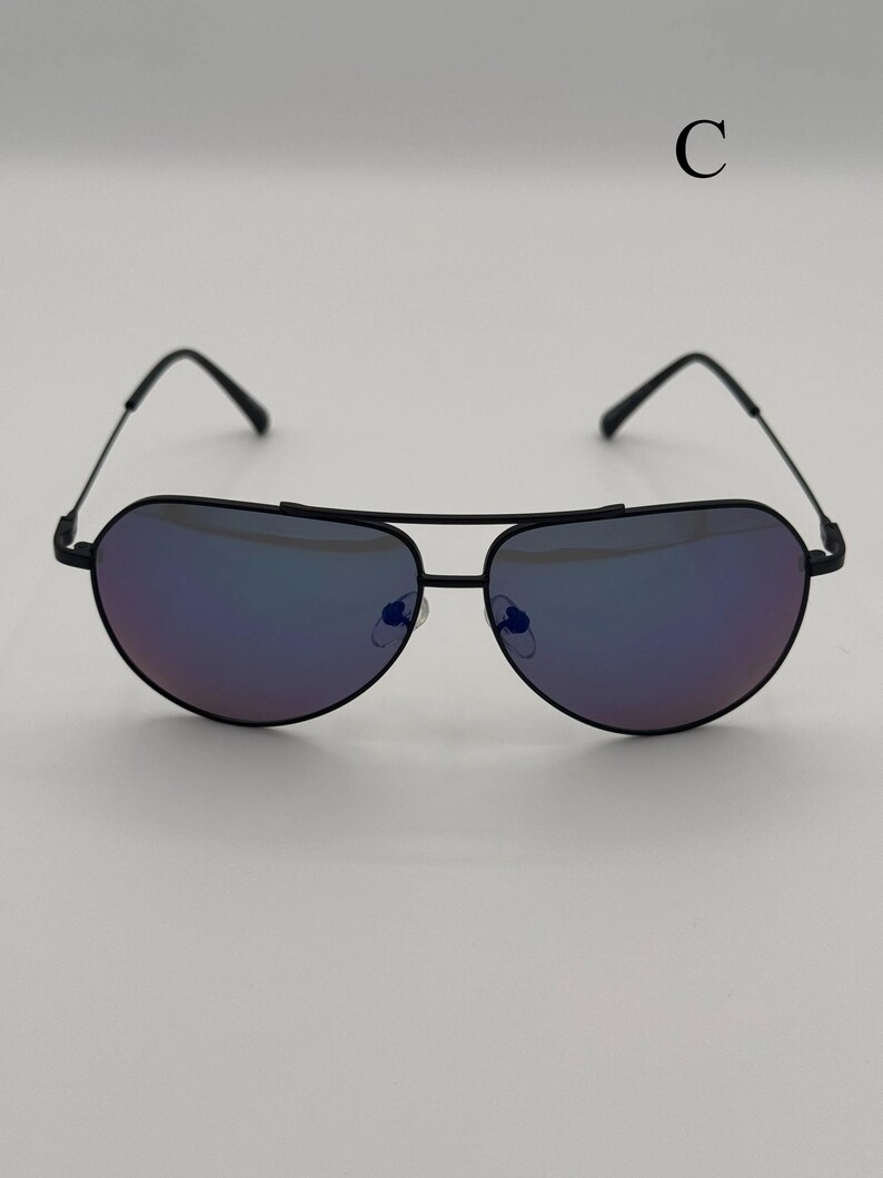 Puede incluir: Gafas de sol de aviador negras con lentes tintadas de p&uacute;rpura. Las gafas de sol tienen una forma cl&aacute;sica de l&aacute;grima y una montura de metal negro. La letra "C" es visible en la esquina superior derecha.