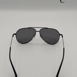 Puede incluir: Gafas de sol aviador negras con lentes de color gris oscuro. Las gafas de sol tienen una fina montura de metal negro y un doble puente. Las patillas son finas y se curvan hacia los extremos. La letra "C" est&aacute; en la esquina superior derecha.