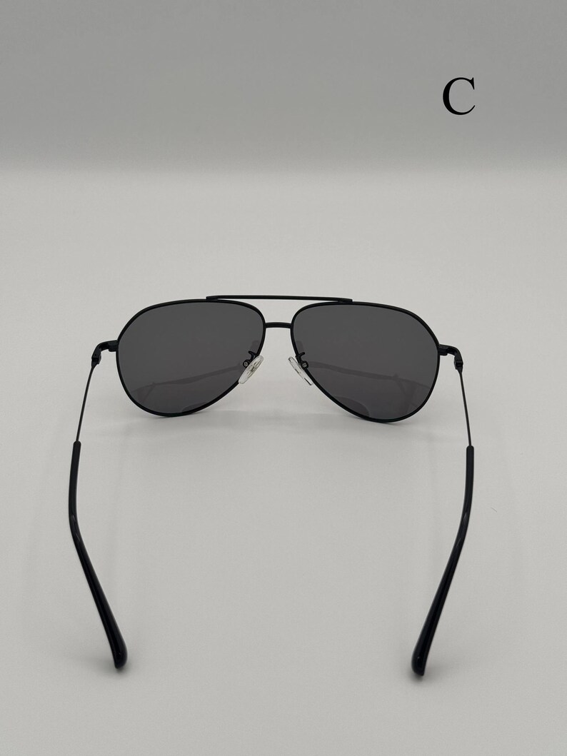 Puede incluir: Gafas de sol aviador negras con lentes de color gris oscuro. Las gafas de sol tienen una fina montura de metal negro y un doble puente. Las patillas son finas y se curvan hacia los extremos. La letra "C" est&aacute; en la esquina superior derecha.