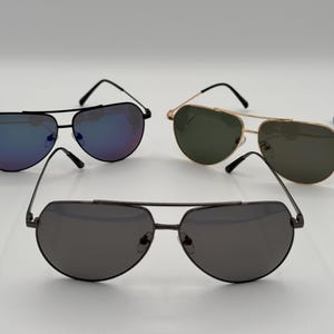Puede incluir: Tres pares de gafas de sol de aviador con lentes de diferentes colores. Un par tiene monturas negras y lentes azul-p&uacute;rpura, otro tiene monturas doradas y lentes verdes, y el tercero tiene monturas gris oscuro y lentes grises.