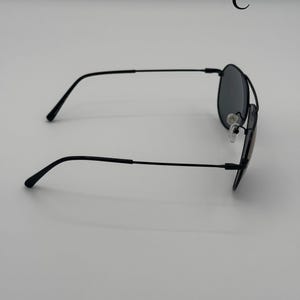 Puede incluir: Gafas de sol negras estilo aviador con lentes oscuras. Las gafas tienen una montura fina de metal negro y almohadillas nasales. La letra "C" es visible en la esquina superior derecha.