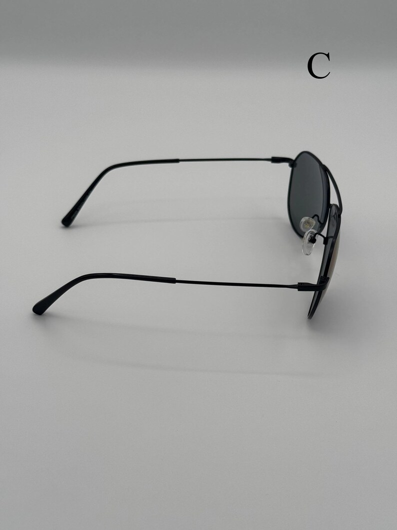 Puede incluir: Gafas de sol negras estilo aviador con lentes oscuras. Las gafas tienen una montura fina de metal negro y almohadillas nasales. La letra "C" es visible en la esquina superior derecha.