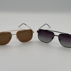 May include: Two pairs of aviator sunglasses. One pair has a gold frame with brown lenses, and the other has a black frame with gradient gray lenses. Both pairs have a double bridge and thin temples.