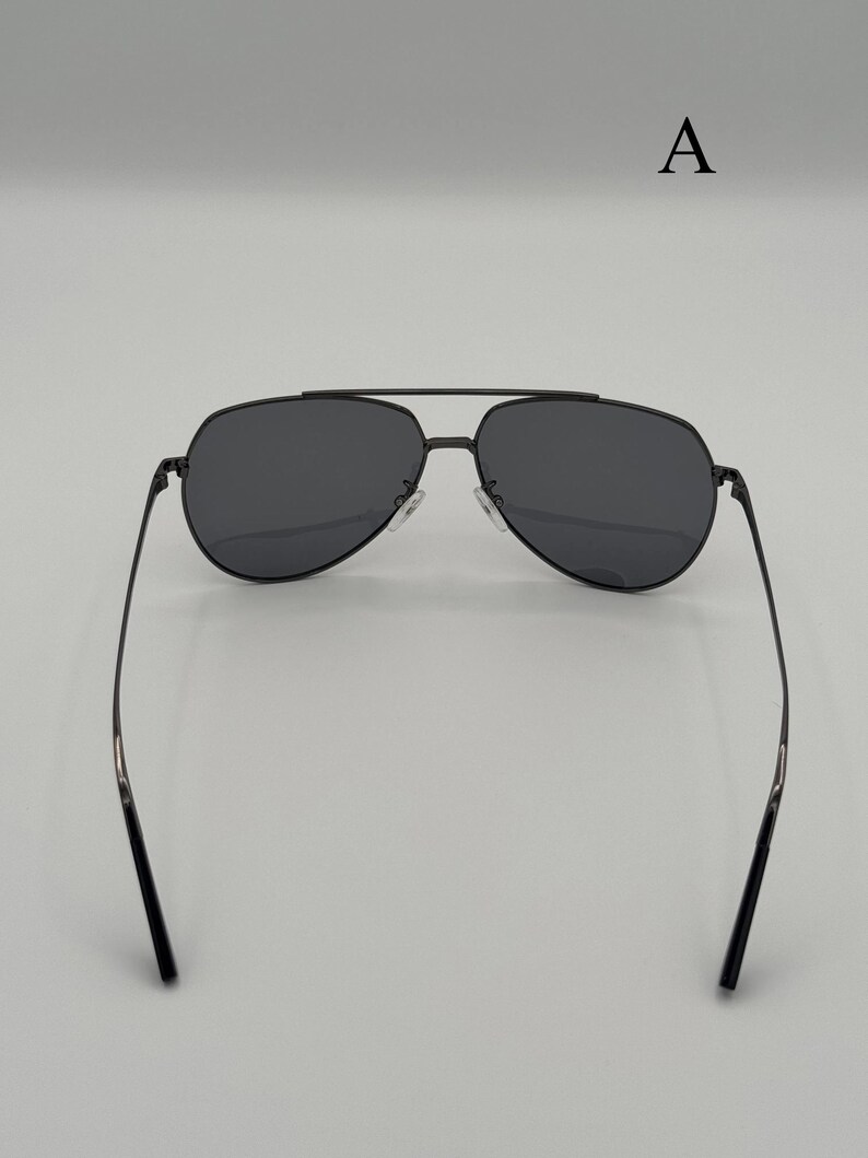 Puede incluir: Gafas de sol de aviador negras con lentes oscuras y una fina montura met&aacute;lica. Las gafas tienen un doble puente y una forma cl&aacute;sica de l&aacute;grima. Las patillas son delgadas y se extienden hacia los extremos. La letra "A" es visible en el fondo.
