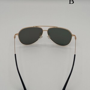 Puede incluir: Gafas de sol de aviador con montura dorada y lentes oscuras. Las gafas tienen un doble puente y patillas negras. La imagen est&aacute; sobre un fondo blanco.